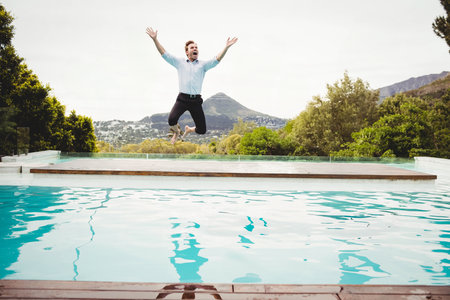 Man wearing blue shirt leaping off wooden deck beside turquoise pool raising arms toward peak. Freedom, adventure, leisure, outdoor, scenic, active, lifestyleの写真素材