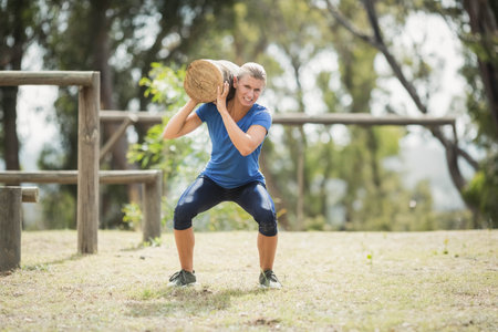 Woman holding wooden log on right-shoulder squatting in grassy area with posts, rails in blue shirt. Outdoor, fitness, training, strength, endurance, obstacle, parkの写真素材
