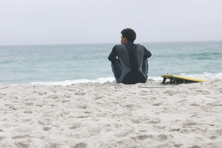 African American man sitting on beach in dark wetsuit facing ocean near yellow surfboard leash. Neoprene, sea, coastline, horizon, solitude, pensive, mutedの写真素材