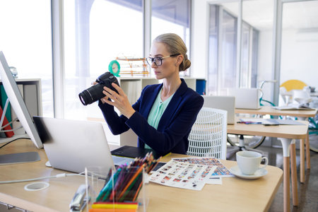 Female creative wearing navy blazer examining DSLR camera, sitting in office with laptop, prints. Photographer, gear, daylight, openplan, modern, bright, airyの写真素材