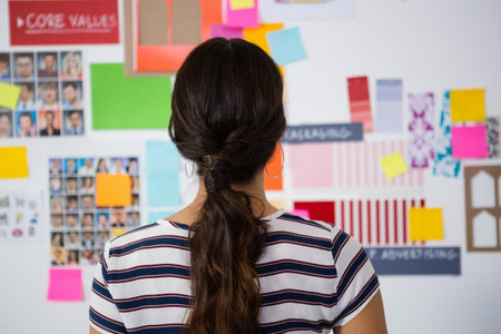 Adult woman standing studying moodboard in studio, wearing tee, reviewing swatches and portraits. Design, palette, color, notes, planning, inspiration, workspaceの写真素材