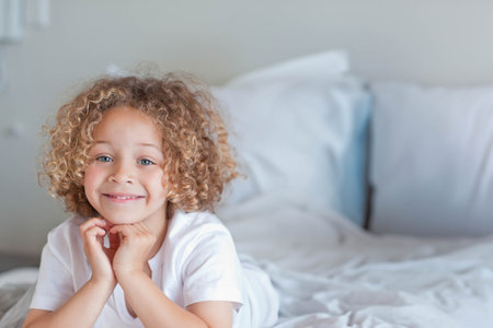 Male child lying on bed wearing white T-shirt, propping chin with hands, looking at camera. Kid, bedroom, pillow, bedding, headboard, softlight, curlyの写真素材