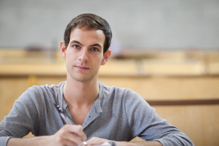 Adult male wearing grey henley sitting at lecture desk, holding pen and writing notes. Classroom, university, student, notebook, paper, wood, benchesの写真素材