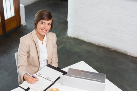 Woman wearing beige blazer sitting at white desk writing in planner with pen, smiling toward camera. Office, workspace, minimalist, modern, professional, focus, productivityの写真素材