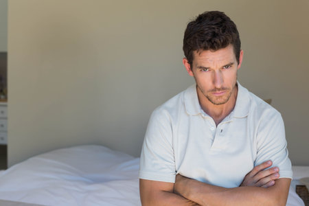 Man sitting on bed edge in bedroom wearing light polo, crossing arms, copy space. Minimalist, neutral, solitary, serious, contemplative, bedding, pillowの写真素材