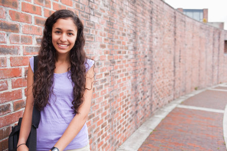 Woman in mid-20s standing, leaning on red brick wall by curved walkway, backpack strap, copy space. Outdoor, urban, student, casual, lavender, pastel, smileの写真素材