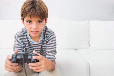Child boy sitting at home on sofa cushions holding black wired controller wearing striped shirt. Gaming, gamer, livingroom, couch, gamepad, minimalist, brightの写真素材