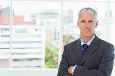 Senior man in dark gray suit, navy tie standing by floor-to-ceiling window at office, copy space. Executive, headshots, professional, business, urban, highrise, skylineの写真素材