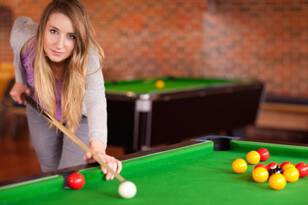 Woman in twenties leaning over pool table in pool hall, aiming cue at white cue ball. Billiards, recreation, casual, brickwall, woodenfloor, cozy, bokehの写真素材