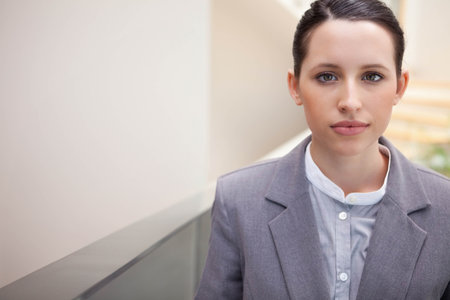 Businesswoman standing by glass railing in office lobby wearing grey blazer and blouse, copy space. Professional, corporate, hallway, contemporary, minimalist, poised, confidentの写真素材