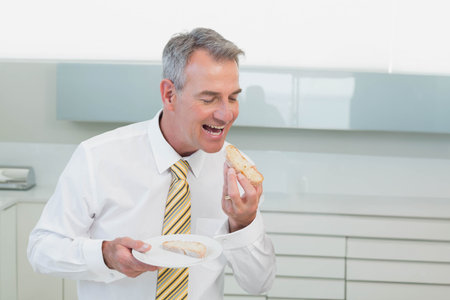 Mature man standing in kitchen wearing white shirt striped tie holding plate with sandwich. Professional, modern, bright, minimalist, workspace, bite, snackの写真素材