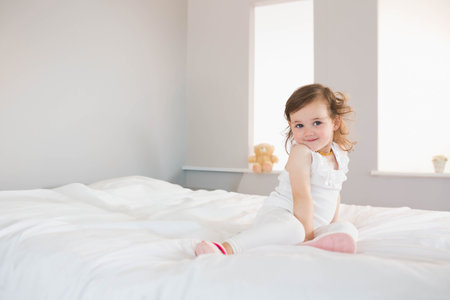 Female toddler sitting on white bed in bedroom with teddy bear wearing white top, copy space. Child, cozy, daylight, neutral, minimalist, pillow, beddingの写真素材