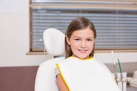 School-age girl sitting in dental chair wearing white bib with yellow trim, dental tray nearby. Pediatric, clinic, dentistry, calm, neutral, instruments, windowの写真素材