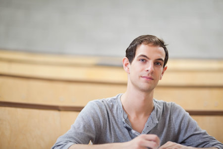 Adult male student sitting in lecture hall with wooden desks, taking notes with pen, wearing henley. Classroom, focus, attentive, writing, notebook, paper, studyの写真素材