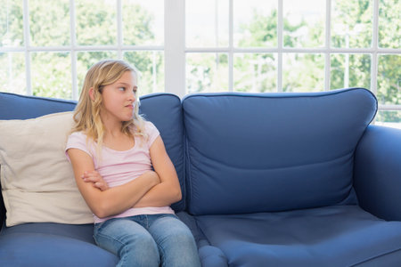 Female child sitting with crossed arms on deep-blue sofa by multi-pane window showing greenery. Girl, pose, cozy, sunlit, bright, cushions, foliageの写真素材