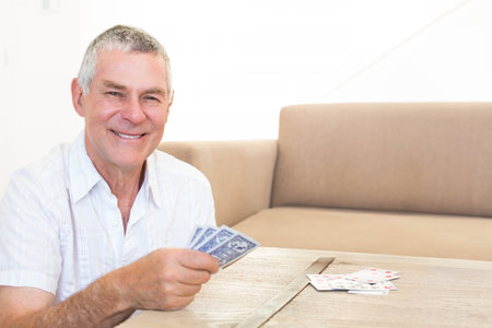 Senior man sitting at wooden table holding playing cards with beige sofa behind, copy space. Elderly, gentleman, retiree, lounge, home, minimal, calmの写真素材