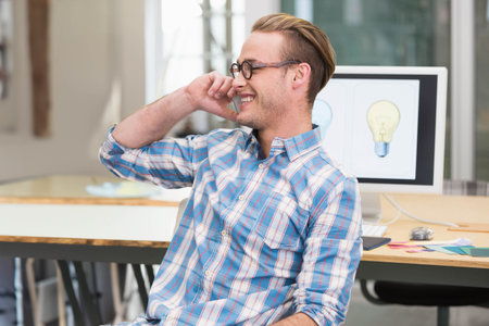 Man phoning, smiling, sitting at wood desk in open office with monitor lightbulbs and swatches. Workspace, designer, glasses, modern, daylight, palette, creativeの写真素材
