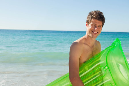 Mid-adult man standing in surf at sunny beach holding green inflatable float shirtless, copy space. Adult, shore, airbed, turquoise, coastal, outdoor, lifestyleの写真素材