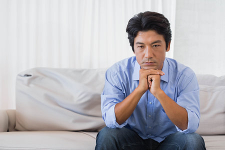 Asian mid-adult man sitting on sofa in living room with white curtains wearing blue shirt, jeans. Interior, home, diffused, minimalist, contemplativeの写真素材