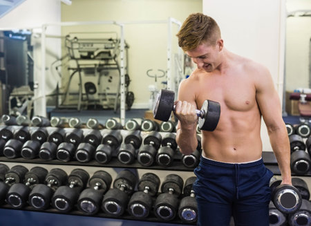 Shirtless man mid-20s doing biceps curl wearing navy shorts by dumbbell rack and mirror, copy space. Gym, weights, fitness, training, strength, focus, dynamicの写真素材