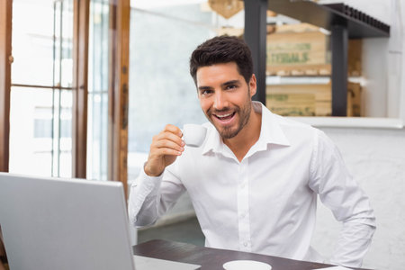 Man in late twenties wearing white shirt holding espresso cup and working on laptop at cafe. Professional, remote, freelancer, coffeeshop, computer, coffee, daylightの写真素材