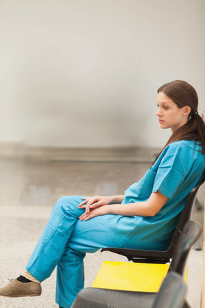Female nurse sitting in clinic wearing teal scrubs on row chair near yellow seat, copy space. Healthcare, uniform, corridor, waiting, serene, polished, minimalistの写真素材