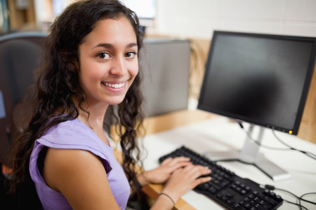 Female teen sitting at lab typing on keyboard, monitor and mouse, wearing lavender top, copy space. Woman, workstation, desk, classroom, education, technology, learningの写真素材