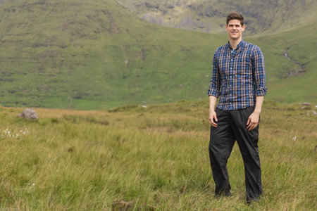 Man standing in valley wearing plaid shirt and dark pants among tall grass, copy space. Outdoor, landscape, nature, remote, hills, grassland, solitudeの写真素材