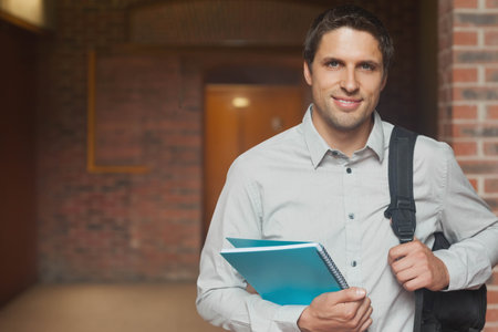 Mid-adult man standing on right in brick corridor, holding blue notebook, gripping backpack strap. Hallway, interior, urban, student, casual, spiralbound, warmthの写真素材