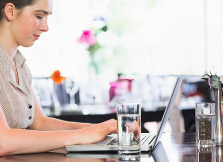 Woman sitting left at reflective cafe table typing on laptop near water glasses, copy space. Cafe, daylight, bokeh, flowers, glassware, productivity, freelancerの写真素材