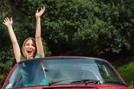 Woman wearing teal short-sleeve top standing through open roof of red convertible, laughing by road. Outdoors, nature, forest, trees, sunlight, vibrant, energeticの写真素材