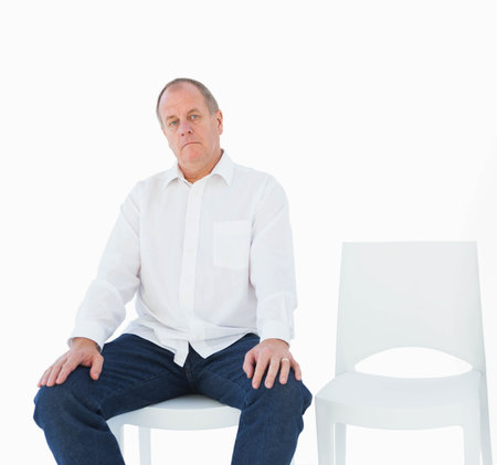 Senior man sitting on white chair in studio, empty chair at right, wearing white shirt, jeans. Mature, adult, seated, stool, seat, minimalist, highkeyの写真素材