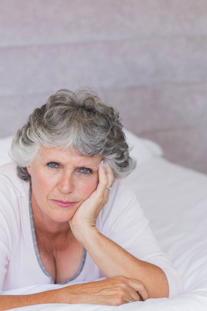 Senior female reclining on bed with white sheets and pillows, wearing pale pink top, showing ring. Elderly, portrait, contemplative, serene, cozy, home, interiorの写真素材