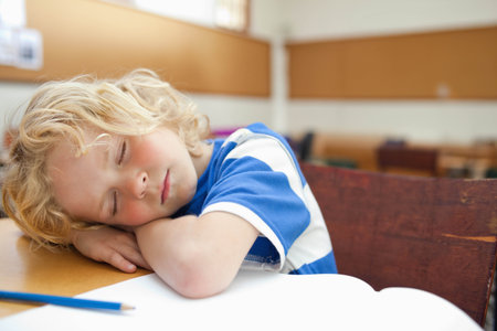 Child boy sleeping on folded arms on wooden desk near open notebook, blue pencil in classroom. Nap, pupil, table, daylight, warmth, bokeh, calmの写真素材