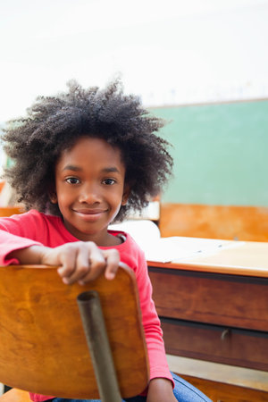 African American child sitting at school desk holding wooden chair back wearing red top blue jeans. Classroom, student, chalkboard, papers, daylight, calm, stockの写真素材