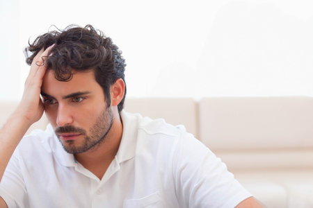 Man sitting on beige sofa at home wearing white polo, holding head and looking down. Sunlight, lounge, cushionsの写真素材
