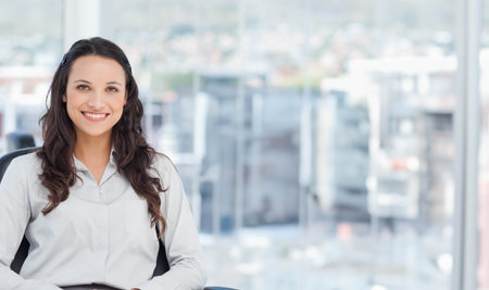 Woman sitting in office chair by large glass window wearing light blouse, smiling, copy space. Businesswoman, portrait, daylight, urban, skyline, modern, cleanの写真素材