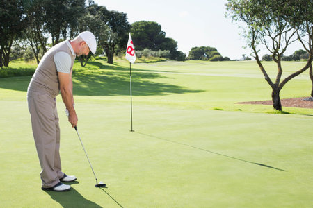 Mature man preparing to putt on putting green, holding putter and eyeing golf ball. Golfer, fairway, flagstick, shadows, sunlight, focus, outdoorの写真素材