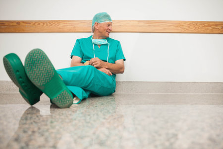 Senior male surgeon sitting in hospital hall leaning on wall by rail wearing scrubs mask clogs. Clinician, corridor, terrazzo, handrail, cap, healthcare, calmの写真素材