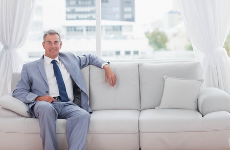 Middle-aged man sitting on pale gray sofa by large window wearing suit and wristwatch. Minimalist, contemporary, urban, cityscape, daylight, glass, loungeの写真素材