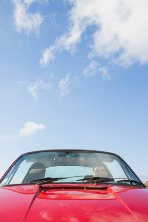 Male driver sitting behind windshield driving red convertible under blue sky with steering wheel. Vehicle, car, outdoors, road, horizon, sunshine, travelの写真素材