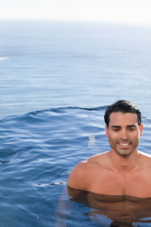 Man floating chest-deep in calm blue water near horizon, sunlit reflections, shirtless, copy space. Outdoors, ocean, pool, adult, serene, bright, relaxationの写真素材