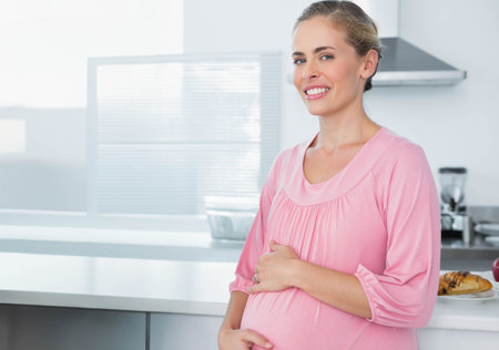 Pregnant woman standing at kitchen countertop in pink top holding belly by pastry plate, copy space. Maternal, bright, modern, minimalist, breakfast, daylight, whiteの写真素材