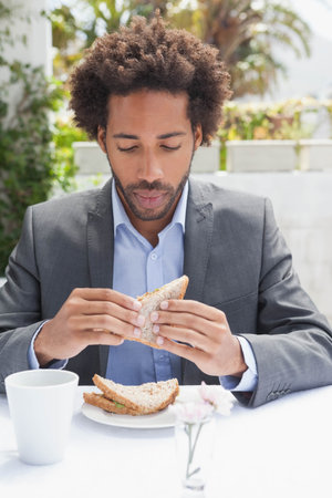 Mid-adult African American man in suit jacket sitting at patio holding seeded sandwich, mug, vase. Businessattire, outdoor, courtyard, cafe, naturallight, sunshine, greeneryの写真素材