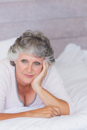 Senior woman reclining on bed, resting left elbow, gazing softly, wearing pale pink top and ring. Portrait, bedroom, pastel, contemplative, intimate, diffused, pillowsの写真素材