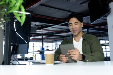 Hispanic man holding tablet sitting at office desk with coffee, monitor, laptop under hanging plant. Technology, workspace, modernelegance, productivity, corporate, freelance, urbanの写真素材