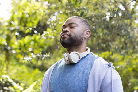 Man standing in sunny park wearing grey hoodie with white headphones at neck. Nature, relaxation, serenity, wellness, outdoor, mindfulness, leisureの写真素材