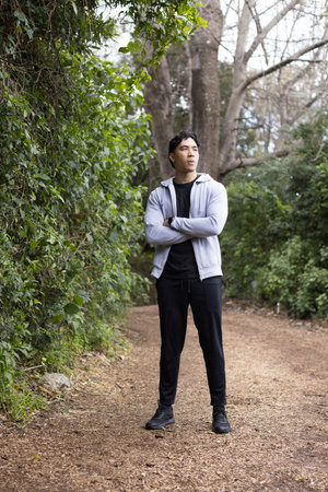 Asian man in athletic attire standing with arms crossed, looking into distance on trail. Nature, adventure, exploration, outdoor, mindfulness, athletic, tranquilityの写真素材