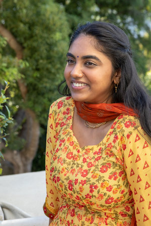 Indian woman sitting on white bench in garden wearing yellow floral kurti and orange scarf....