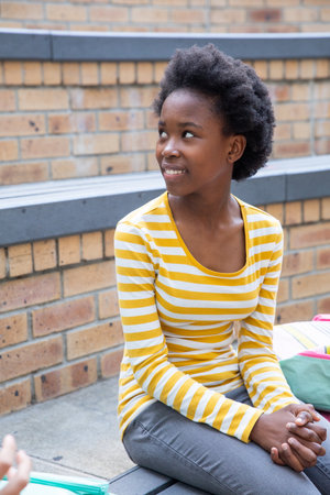 African American teenage girl sitting on school steps against brick wall with green backpack. Youth,...